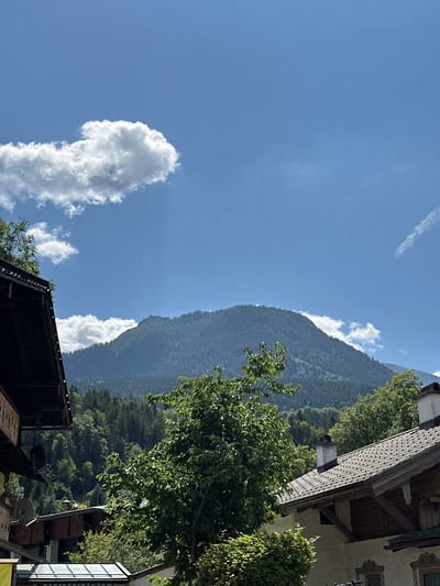 This is a photograph of the Watzmann mountain in Berchtesgaden, Germany, taken from a village street. The mountain is covered in lush green trees and has a large, imposing presence. The sky is a vibrant blue with a few fluffy white clouds scattered across it. In the foreground, there are several traditional Bavarian houses with their characteristic wooden balconies and tiled roofs. A large green tree with full foliage is in the middle ground, obscuring part of the view of the mountain. The overall impression is one of a picturesque mountain village on a sunny day.