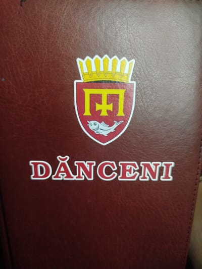 The image shows a close-up of a maroon, textured surface, possibly leather, bearing a coat of arms and text. The coat of arms is a shield with a crown at the top. Inside the shield, there is a stylized yellow letter "F" and a white fish. Below the coat of arms, the text "DĂNCENI" is prominently displayed in large, white, stylized letters with a red outline. The text and emblem appear to be a logo or insignia. The background is a plain, dark maroon color with visible texture. The overall impression is of a decorative element on a notebook, book cover, or similar item. The location in Nimoreni, Moldova, is not visually discernible from the image itself.