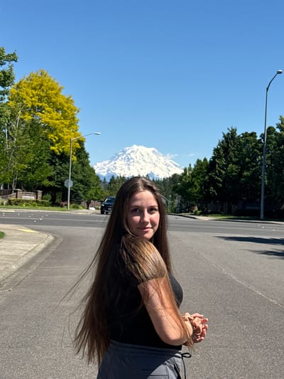 A young woman with long brown hair and a black t-shirt stands on a paved road in South Hill, USA, looking back towards the camera. Behind her, a snow-capped mountain, possibly Mount Rainier, rises majestically against a clear blue sky. Lush green trees line the street, with a bright yellow tree adding a splash of color to the scene. A street lamp and a stop sign are visible, along with a black car in the background. The woman is holding her hands clasped in front of her. The image captures a sunny day with ample daylight.