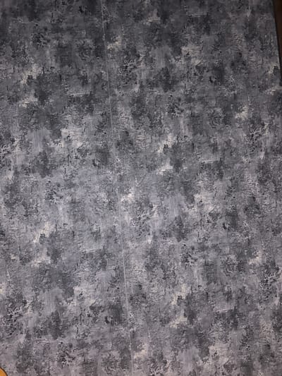 This is a top-down, full-frame shot of a textured, abstract patterned surface, likely wallpaper or a painted wall. The pattern consists of irregular splotches and strokes in various shades of gray, black, and white, creating a mottled or distressed effect. A thin, light-colored vertical line, possibly a seam or a crack, runs down the center of the image from top to bottom. There is no indication of Apsheronsk, Russia, or any other specific location, people, actions, or interactions. The image is purely a visual representation of a textured surface.