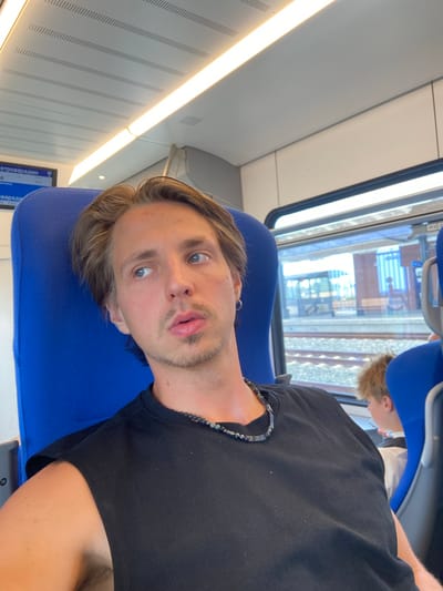 A man with blond hair, light blue eyes, and a goatee sits on a train, looking to the left. He is wearing a black sleeveless t-shirt and a beaded necklace. A blue train seat is visible behind him and to his left. Through the window to his right, a train station platform can be seen, with another passenger sitting in the background. The train appears to be in motion, as evidenced by the blur in the background. The man is in the city of Indjija Municipality, Serbia.