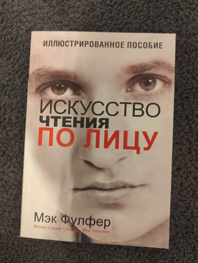 This is a top-down, eye-level, full-shot photograph of the cover of a book titled "ИЛЛЮСТРИРОВАННОЕ ПОСОБИЕ ИСКУССТВО ЧТЕНИЯ ПО ЛИЦУ" by Мэк Фулфер. The book cover features a black and white, close-up image of a person's face, with their eyes, nose, and mouth prominently displayed. The title is overlaid on the face in large, bold letters. The author's name and a subtitle, "Иллюстрации Сандры Мур Уильямс," are printed in smaller text at the bottom of the cover. The book is lying on a dark, textured surface, possibly a carpet or fabric. There is no discernible location in the city of Schwäbisch Gmünd, Germany visible in the image. The image is focused on the book, and the background is out of focus. There are no people or actions depicted other than the static presence of the book.