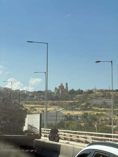 The image shows a view of Polichni Municipal Unit, Greece, on a clear day. In the foreground, a truck is parked on the side of a road, loaded with construction materials. A person is seen riding a motorcycle behind the truck. In the background, a domed building, possibly a church, is visible on a hillside, with houses and vegetation surrounding it. Several streetlights are also present in the scene. The sky is blue with some white clouds. The image appears to be taken from a vehicle moving along the road.