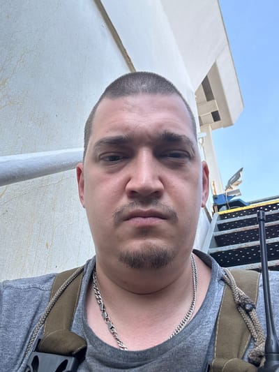 A close-up, eye-level, outdoor selfie shows a man with short hair, a mustache, and a goatee. He is wearing a grey t-shirt, a silver chain necklace, and a brown backpack with straps. He is holding a black walkie-talkie in his right hand. In the background, there is a white wall, a metal railing, metal stairs, and a blue sky. The scene appears to be on a ship or a structure with industrial elements. There are no discernible landmarks that would place the image in Uribia, Colombia.