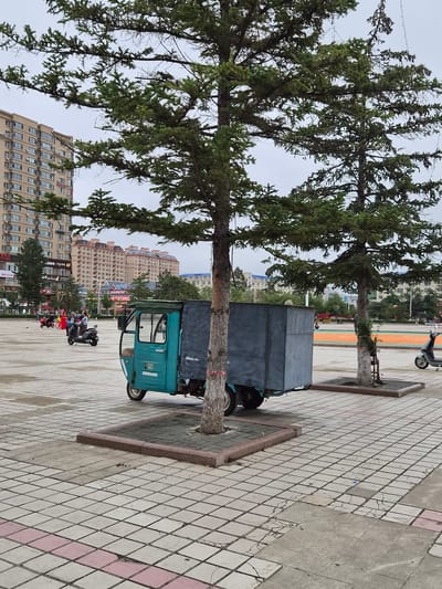 The image shows a paved square in the city of Hunchun, China, with buildings in the background. In the foreground, a turquoise three-wheeled vehicle with a large, gray cargo box is parked near a tree. The vehicle has a covered cab and appears to be a delivery or utility truck. A motorcycle is parked to the right of the vehicle, and two people are riding another motorcycle in the middle ground. In the far background, there are several multi-story buildings, some with balconies and air conditioning units. The sky is overcast, and the square is sparsely populated, suggesting it is either early morning or late afternoon. The pavement is tiled with a pattern of gray and pink squares, some of which appear worn or cracked. The overall scene depicts a typical urban environment in Hunchun, likely during a period of low activity.