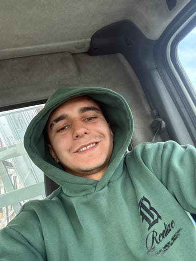 A male individual is photographed in a close-up, selfie-style shot from within a vehicle in Mlada Gvardia, Bulgaria. He is positioned centrally, looking directly at the camera with a smile, revealing his upper teeth, which exhibit a minor diastema between the central incisors. He has light brown eyes, dark eyebrows, and visible stubble on his chin and upper lip. He is wearing a teal-green hooded sweatshirt with the hood pulled over his head. The sweatshirt features a black graphic on the left chest area: a stylized Gothic letter, interpreted as a 'B' or 'R', positioned above the script word "Realize". Below this, partially legible text states: "TRUST NOBODY - I AM NOTHING LOYALTY AND STRENGTH". The vehicle's interior shows a light grey headliner and upper pillar trim. A dark grey plastic trim piece and a seatbelt mechanism are visible on the right side of the frame. Through the vehicle's left window, blurred vertical structures of a light color are discernible, while a segment of a blue sky with white clouds is visible through the upper right portion of the frame.