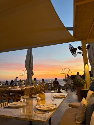 The image captures an outdoor restaurant terrace in Palaiokastritsa, Greece, during sunset. The sky, visible above the horizon, displays gradients of orange, pink, and light blue, transitioning to a deeper blue at the top. The Mediterranean Sea is visible in the background.

The dining area is partially covered by large, light yellow fabric shade sails. A long, light-colored rectangular table, likely granite or marble, occupies the foreground, set for two with light-toned plates, silverware (fork and knife), and inverted clear glasses. A gold-colored decorative object or lamp stands between the place settings. To the immediate right, a section of a cushioned bench seat with light-colored pillows is visible.

In the midground, approximately 10-15 individuals are seated at various round and rectangular tables, or standing. Most tables are equipped with wicker-backed chairs. One person, dressed in a white shirt and dark trousers, presumed to be a server, stands near a table on the far left. A large, collapsed, cream-colored patio umbrella is centrally positioned within the dining space. Decorative arched lighting fixtures are present among the tables. A yellow structural column on the right supports a black wall-mounted industrial fan and two spotlights. Potted plants, including what appear to be small olive trees, are interspersed throughout the terrace. Patrons are engaged in dining and social interaction under the ambient light of the sunset and subtle artificial illumination.