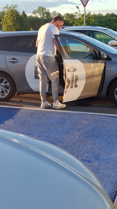 An adult male wearing a white t-shirt and grey sweatpants stands beside a dark grey hybrid car with the driver's side door open. The car has a large white circular logo with a black "L" shape and the word "bolt" in black text on the door. The man is leaning into the car, with his right hand near the interior door handle. He is wearing white socks and light-colored athletic shoes. The scene appears to be a parking lot, with a blue-patterned asphalt surface in the foreground. In the background, there are trees and a clear sky with a few clouds. A triangular white and red sign is visible in the upper right corner of the image, possibly indicating a traffic warning. The overall lighting suggests it is late afternoon or early evening. The location is Tiskre, Estonia.