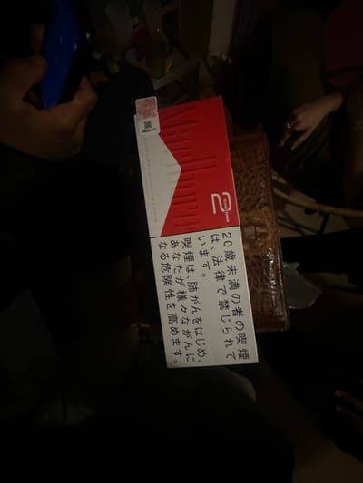 The image shows a pack of Marlboro cigarettes on top of a brown, textured box, possibly a wallet or a tissue box. The cigarette pack has Japanese warning labels printed on its side. A hand holding a smartphone is visible in the lower left corner of the image. The scene appears to be indoors with multiple people present. There is nothing in the image to indicate that it's in Montreuil, France.