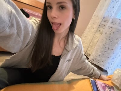 The scene is an indoor setting in a private residence located in the city of Волховец, Russia. A young adult female with long, dark hair and fair skin is depicted in a low-angle selfie. She is wearing a light grey zippered hoodie over a black top and has small earrings. Her expression is characterized by an extended tongue. Her left arm is raised, holding the camera, while her right hand rests on an orange surface. In the immediate foreground, on this orange surface, lies an open magazine or book featuring anime-style imagery, next to a crinkled white plastic bag. The background on the left shows a wooden furniture item, possibly a bed frame or headboard, with folded pink and black fabric. To the right, a window is visible, covered by a white sheer curtain with a textured pattern, through which natural light enters the room.