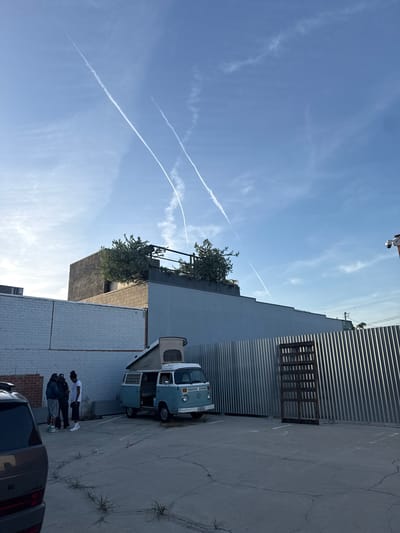 The image shows a partly cloudy sky over a parking lot in what appears to be an urban area in Culver City, California. Three visible contrails bisect the clear blue sky. A blue and white vintage Volkswagen van is parked adjacent to a corrugated metal wall and a white brick building. Three people stand near the van. The rear of a dark-colored minivan is visible at the bottom of the image. A tall gray building is adjacent to the VW. Plants grow on the roof of the tall building.