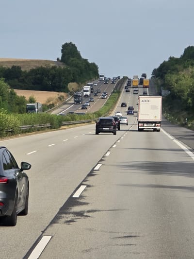 The image depicts a busy highway in the vicinity of Brechen, Germany, with a significant volume of traffic. In the foreground, a dark-colored car is partially visible on the left side of the frame, indicating the perspective is from within another vehicle traveling on the highway. The highway stretches into the distance, curving and ascending a hill.
Multiple lanes of traffic are present, with various vehicles including cars, trucks, and a semi-trailer truck with "ASS Transports" branding. Some vehicles are in motion, while others appear to be part of a traffic queue, particularly further up the incline. The highway is flanked by green vegetation and trees on both sides. A field of dry, golden grass is visible on the left side of the highway, contrasting with the lush greenery. The sky is overcast with a pale, hazy blue. The overall scene captures a typical highway scene with heavy traffic flow under a diffused sky.