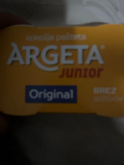 The image displays a close-up, slightly out-of-focus view of a packaged food product. The primary subject is a yellow-orange rectangular container, held by a human hand, partially visible on the left. The container's label features prominent white lettering for the brand "ARGETA," with the word "JUNIOR" in red positioned directly below it. Above the "ARGETA" logo, the text "Kokošja pašteta" (Chicken pâté) is visible in a lighter yellow hue. A blue rectangular label on the lower-left portion of the package identifies the product variant as "Original." To the right of the "Original" label, the words "BREZ aditivov" (without additives) are partially visible. No other individuals or actions are discernible. The background is dark and highly blurred, providing no contextual details about the surrounding environment or its specific location. The image itself provides no visual information to determine its specific location within the city of Brežice, Slovenia.