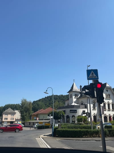 The image captures a street view in Bridel, Luxembourg, on a clear, sunny day. In the foreground, a red car is seen driving on a paved road. A traffic light with a red light illuminated is visible on the right side of the image, adjacent to a pedestrian crossing sign. Behind the traffic light, a white building with distinctive architecture, featuring turrets and arched windows, stands prominently. A sign on this building reads "BRECK". Further back, other buildings, trees, and a lush green hillside form the backdrop under a vast blue sky. The overall scene depicts a tranquil urban environment with clear visibility.