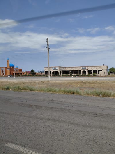 The image shows a street scene in the city of Бұлақты, Kazakhstan. On the left side of the image is a mosque with orange walls and blue domes. A white car is parked in front of the mosque. To the right of the mosque is a long, beige building with arched windows and a covered walkway. The building appears to be a commercial establishment, with signage in an unidentified script visible above the windows. A power line pole with wires runs through the middle of the image, obscuring parts of the sky and the buildings. The foreground of the image is an asphalt road with visible cracks and patches, suggesting wear and tear. Dry grass and shrubs are present on the median and along the sides of the road. The sky is blue with scattered white clouds and a dark shadow from an unseen object is visible across the top of the frame. The overall impression is that of a functional, somewhat utilitarian area with signs of development and ongoing infrastructure.