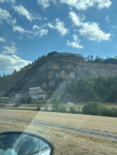 The image depicts a landscape scene with a large quarry or mining operation on a mountainside. In the foreground, a dry, golden field stretches across the frame, with a dark dirt path cutting through it. A car's side mirror is visible in the bottom left corner, reflecting a person's face.

Further back, a complex of industrial buildings, likely part of the quarry operation, is situated at the base of the mountain. These structures are made of concrete and metal, with conveyors and other equipment visible. Trees and vegetation surround the buildings and climb up the lower slopes of the mountain.

The mountain itself is a prominent feature, with a large, scarred face indicating quarrying activities. Layers of rock and soil are exposed, and sparse vegetation grows on some ledges. The top of the mountain is covered with a dense forest of evergreen trees.

A white camper van is parked on a road in the middle ground, near the industrial buildings. The sky above is a bright blue with scattered white clouds, suggesting a sunny day.

The location is identified as the quarry in the city of Haslach im Kinzigtal, Germany.