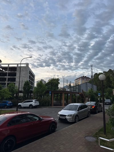 A low-angle, full outdoor shot captures a street scene in the Vardane Microdistrict, Russia, under a cloudy sky. In the foreground, a dark red Mercedes-Benz sedan is parked on the right side of a brick sidewalk. To its left, a silver Ford Focus hatchback is parked. Further down the street, a dark-colored sedan is also parked. Across the street, a white SUV is parked in a lot. Behind the parked cars, a large, unfinished concrete building stands tall, with exposed floors and structural elements. Beyond that, trees line the street and a fenced sports court is visible. In the background, a multi-story residential building with a reddish-brown roof and several stories of windows is visible, with a tall communications tower to its right. A white spherical lamp post stands near the sidewalk. The overall impression is of an urban environment with a mix of residential and commercial structures, under a partly cloudy sky.