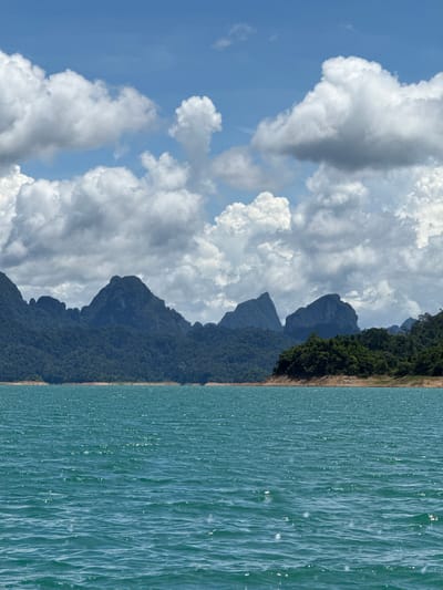 This is a landscape photograph taken from a body of water, likely in or around Ban Chiao Lan, Thailand. The foreground is dominated by a vast expanse of turquoise water, rippling with gentle waves that reflect the sunlight, creating a shimmering effect. The middle ground features a densely forested shoreline transitioning into a series of dark, rugged mountains that rise sharply against the horizon. These mountains are covered in thick vegetation, with some peaks appearing jagged and rocky. The sky above is a vibrant blue, adorned with a dramatic display of fluffy, white cumulus clouds, some of which are tinged with grey, suggesting an impending change in weather or the presence of shadow. The overall scene evokes a sense of natural beauty and tranquility, characteristic of a tropical lake or reservoir setting.