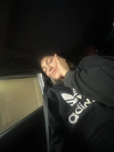 The scene is located within a vehicle in Airolo, Switzerland. A female individual occupies the foreground, appearing to be in a passenger seat, secured by a visible seatbelt across her torso. She has closed eyes and a slight smile, with her right hand resting against her right cheek. A silver-colored ring is present on her right ring finger. She wears a black hooded sweatshirt featuring a white Adidas Trefoil logo. Her face is brightly illuminated by a warm-toned external light source originating from the left side of the vehicle. A second, indistinct individual is partially visible in the blurry, dimly lit background. The vehicle's interior is dark, with a window on the left revealing a blurry, yellowish-beige exterior environment.