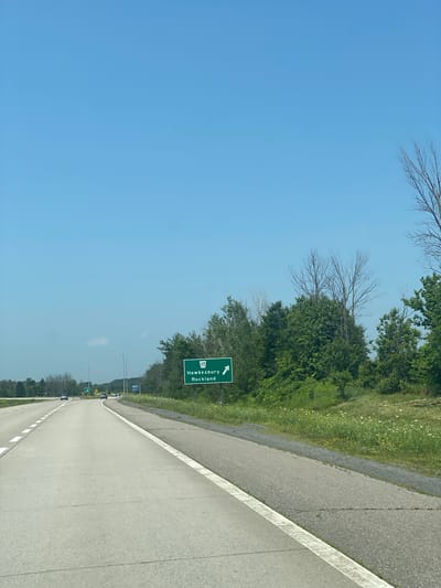 A highway sign on the right side of a highway indicates that Route 17, leading to Hawkesbury and Rockland, is to the right. The sky is clear and blue. The highway has two lanes of traffic, with a white line marking the right shoulder. There are several cars on the highway in the distance. The right side of the highway is lined with trees and green grass. The scene appears to be in East Hawkesbury, Canada.