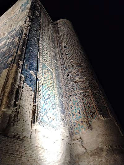 The media file displays a low-angle, wide shot of a large, ornate building, possibly a mausoleum or mosque, at night in Shahrisabz City, Uzbekistan. The building's facade is covered in intricate geometric tilework, primarily in shades of blue, turquoise, and white. The tiles form complex patterns, including repeating diamond shapes and what appear to be Arabic inscriptions.

The structure is massive, with visible brickwork at its base, some of which is crumbling and aged. Large sections of the tilework have fallen away, revealing the underlying brick and plaster, giving the building a weathered and ancient appearance. The contrast between the illuminated lower portions and the dark night sky creates a dramatic effect. Artificial lighting illuminates the bottom part of the structure, casting bright lights upwards and highlighting the textures of the brick and the lower tiles.

The perspective from below emphasizes the towering height and grandeur of the building. The scene is static, with no people or activity evident. The time of day is clearly night due to the black sky and artificial lighting. The weather is not discernible. The architectural style and the presence of elaborate tilework are strong indicators of Central Asian or Islamic architecture. There is no visible text that can be clearly read or deciphered from the image.