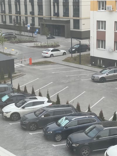 A street view captures a parking lot in front of an apartment building in Sokilnyky, Ukraine. Several cars are parked in designated spots, including a white sedan, a silver wagon, a dark gray SUV, a dark blue wagon, and a black SUV. The apartment building features modern architecture with beige and brown exterior colors, and multiple windows. The ground is paved with interlocking bricks. The overall lighting suggests an overcast day.