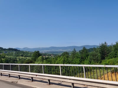 The image depicts a clear blue sky above a landscape viewed from a bridge. In the foreground, a white and black metal guardrail runs horizontally across the frame. The road surface is visible in the lower portion of the image. Beyond the guardrail, rolling green hills are covered with lush trees, with a few buildings and structures scattered throughout. In the distance, hazy mountains form the horizon. The scene appears to be set in the vicinity of Golemo Buchino, Bulgaria, given the distinctive geographical features and vegetation consistent with the region.