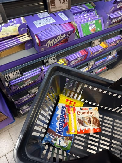 The image shows a shopping basket filled with various chocolate bars and candy, positioned on the floor in front of a shelf display of more chocolate bars in a retail environment. The shelves are stocked with Milka brand chocolates, specifically Milka "Weiße Schokolade" (White Chocolate) and Milka "Bunte Haselnuss" (Mixed Hazelnut). Some of the chocolates have visible price tags indicating "1,82" and "Edeka".

In the shopping basket, there are several items: a yellow packet of "Kinder" brand candy, a blue and brown wrapped "Knoppers" bar, and a "Kinder Country" chocolate bar. A portion of another item with a green background and "GEWINNE" (Win) is also visible.

The background reveals a store aisle with brightly lit shelves and pricing labels common in German supermarkets. The overall lighting suggests an indoor commercial setting. Based on the presence of Edeka branding and the type of products, the scene is likely located in a supermarket in Stadtallendorf, Germany.