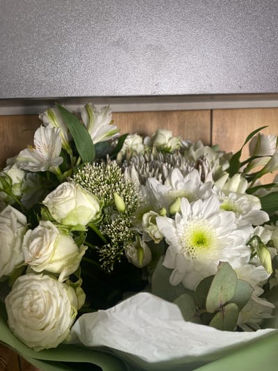 A close-up, eye-level shot captures a bouquet of white flowers, primarily roses and chrysanthemums, with a subtle green hue and hints of light pink and yellow. The flowers are arranged with a variety of textures and shapes, including delicate small white buds. The bouquet is presented against a backdrop of textured wooden planks and a muted gray metallic surface. The composition is centered on the floral arrangement, with some of the stems and wrapping paper peeking out from the bottom. There are no people or animals present in the image. The lighting is soft and diffused, highlighting the details of the flowers. The scene appears to be indoors. The location in Osentsy, Russia is not depicted in the image.