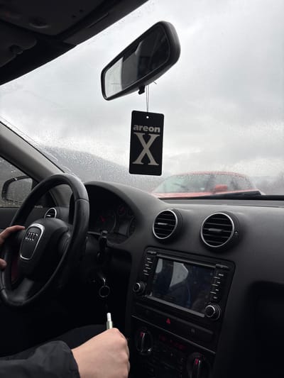 The image captures an interior view from the driver's seat of an Audi vehicle, specifically located in Malinovo, Bulgaria, looking forward through the windshield. Raindrops are visible on the exterior of the windshield, indicating wet or damp weather conditions under an overcast sky. A person's left hand is positioned on the black, multi-function steering wheel bearing the Audi logo. Their right hand holds a white and metallic tubular object, resembling a vape pen, situated near the lower central console area. A set of keys is visible in the ignition. A black rectangular "areon X" air freshener hangs from the rearview mirror, which itself reflects the grey sky. The vehicle's dashboard is black, featuring two circular air vents with chrome rings on the passenger side, and a central infotainment screen displaying what appears to be a navigation interface with "22 km" and "13:34" visible. Below this, a climate control panel shows a temperature reading of "21.5°C". Through the windshield, the blurred red hood and upper portion of another vehicle are visible in the foreground.