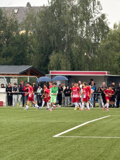 This image depicts an outdoor soccer field scene in Käerjeng, Luxembourg.
The foreground features a green artificial turf pitch with visible white field markings, including segments of the halfway line and a perpendicular line in the lower right. Several young male soccer players are present on the field. Most are wearing red and white striped jerseys with "LEHRMANN" and "SPORT" text, red shorts, and white socks, with brightly colored cleats (rainbow, pink, yellow, blue). One player towards the center-right is distinct in a bright lime green jersey, matching shorts, and white socks, suggesting a different team or a goalkeeper. The players are in various stages of motion, appearing to walk or light jog across the field.
Behind the playing area, a metal barrier separates the field from a crowd of approximately twenty-five spectators of mixed ages and genders, dressed in casual attire. Many are observing the field, with some holding beverages. On the far left, a person holds a bottle. Towards the right, a man in a red cap and beard is visible.
On the left side of the midground, a covered dugout or structure with a dark corrugated metal roof and wooden supports is visible, partially obscured by a goal net. Towards the right, a white and red portable container-like structure, possibly a concession stand or official's booth, stands. Two individuals are partially visible beneath two open blue umbrellas in front of this structure, suggesting rain or sun protection.
The background is dominated by lush green trees with varied foliage. Behind the trees on the left, a dark tiled building roof with a skylight and a chimney is partially discernible, along with hints of a lighter-colored wall. The sky is overcast and uniformly light grey.