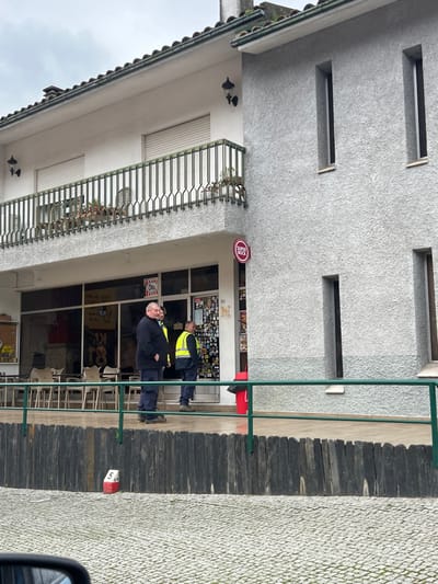 The image depicts the exterior of a two-story building in Santa Marta de Penaguião, Portugal, under an overcast sky. The structure features a commercial ground floor with a patio and a residential or utility upper floor.

The ground floor consists of a storefront with large glass windows and an entrance on the left side, leading to an outdoor patio furnished with several light-colored tables and chairs. A circular red "SUPER BOCK" sign is mounted on the building's exterior to the right of the entrance. A red fire hydrant and a red trash bin are situated near the right side of the storefront entrance. The storefront windows display various items and posters. A small sign displaying "Ola" is visible on one of the glass panels, and the number "71" is above the entrance. The building's right section is a textured grey wall featuring two narrow, vertical windows on the ground level and two identical windows on the upper level.

The upper floor features a white facade with a balcony enclosed by a green metal railing. Two windows with closed white roller shutters are visible behind the railing, accompanied by two wall-mounted lights. The roof is tiled and exhibits visible green moss.

Three adult males are present on the ground-floor patio. The leftmost male, with short grey hair, wears a dark long-sleeved shirt and dark pants, standing with hands in his pockets and facing slightly left while smiling. The middle male, partially obscured, and the rightmost male both wear yellow high-visibility vests over dark garments and dark pants, standing upright.

In the mid-ground, a dark grey/black wooden fence acts as a barrier, topped with a green metal railing. A small white and red marker, numbered "5," is visible on the ground directly in front of the fence. The immediate foreground is paved with grey cobblestones, and a portion of a vehicle's side mirror is visible in the bottom left corner.