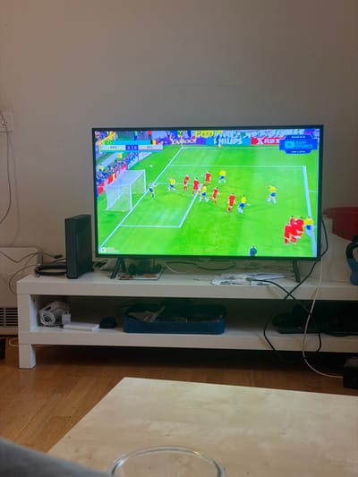 An interior scene captured from a low vantage point reveals a television displaying a soccer match, situated within a residential setting in Adliswil, Switzerland. The television, a flat-screen model, is centrally positioned on a white, two-tiered entertainment console. The screen shows a match between Brazil (BRA) and Belgium (BEL) with a 0-0 score, identified as "ROUND OF 16" and dated "JUNE 3, 2022." Players in yellow/blue and red/yellow kits are visible on a green field, with a goal and a goalkeeper in light blue prominently featured. Logos for "Sian Sports," "Yahoo!," and "Philips" are discernible on the screen.
To the left of the television, a black, rectangular electronic device stands vertically. Various black cables are visible, extending behind and below the television, connecting to components on the stand. The top surface of the console holds indistinct small items. On the lower shelf of the console, from left to right, are a coiled white cable, a white rectangular object, a black computer mouse, and a dark blue rectangular tray containing several items.
The background consists of a light-colored wall. A white electrical outlet with an attached cable is visible on the upper left wall. Below this, a white radiator or heater unit is partially visible. The floor is covered with light brown wooden planks or laminate. In the immediate foreground, a light-colored wooden coffee table is partially visible, with the rim of a clear glass or cup resting on it. A sliver of gray upholstery, likely from a sofa, is visible at the bottom left. A red plastic bucket or container is partially visible on the far right side of the frame.
