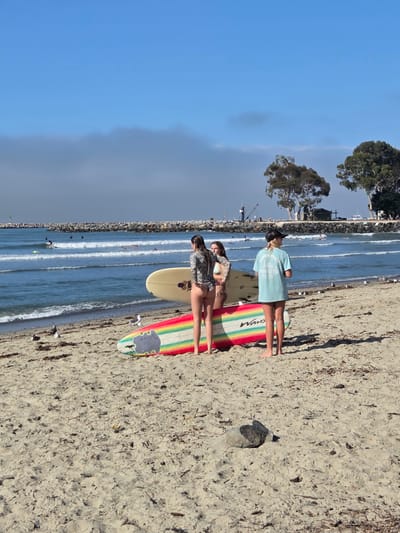 The image depicts a beach scene in Dana Point, California, United States of America. Three young women are present on the sand, two of whom are standing next to surfboards. One surfboard is a light cream color with an American flag design near the tail, while the other is vibrantly colored with rainbow stripes. The woman on the far right is wearing a light blue t-shirt and black baseball cap. The other two women are wearing swimsuits. In the background, the Pacific Ocean is visible with several surfers in the water. A jetty made of large rocks extends into the ocean, and there are trees and some structures on the distant shore. The sand is covered with dried seaweed and debris. Several seagulls are scattered on the beach. The sky is partly cloudy with a band of fog on the horizon.