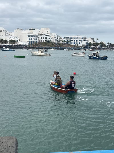 The image shows two people in a small rowboat in the water, likely in the harbor of Arrecife, Spain. The buildings visible on the left of the image are characteristic of the architecture in the city. One man is wearing a basketball jersey with the number 13 and the name "Nash" on the back, and a red hat, while the other appears to be paddling. Several other boats are anchored in the harbor. The water is slightly choppy, and the sky is overcast.