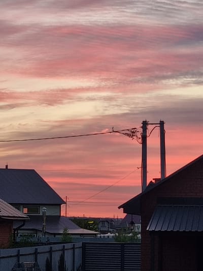 This media file captures a vibrant sunset over a residential area, likely in Iskra, Russia. The sky is dominated by streaks of pink and white clouds, suggesting a dramatic and colorful end to the day. Partially visible buildings, with dark, sloped roofs and some siding, are silhouetted against the brightly lit sky. A utility pole with wires is prominently featured in the foreground, adding an element of the urban or suburban landscape to the natural beauty of the sunset. The overall mood is serene and visually striking, highlighting the transition from day to night. There is no visible text in the image.