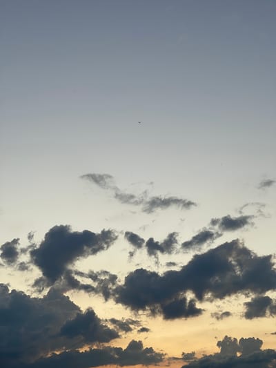 A wide shot captures the sky above Zaķumuiža, Latvia, at what appears to be dusk. The upper portion of the image is a pale, almost pastel blue, transitioning to a soft yellow and orange hue towards the horizon. Dark, cumulus clouds dominate the lower half of the frame, with some lighter, wispy clouds scattered higher up. A single, tiny black silhouette of a bird is visible in the upper center of the image, and another smaller silhouette is to the right of the first. The scene is devoid of any human presence or man-made structures, focusing solely on the atmospheric conditions and the vastness of the sky.