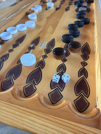 A high-angle, perspective shot shows a backgammon board. The board has a light brown wood finish with dark brown and black decorative markings. There are black checkers arranged in a row on one side of the board, and white checkers arranged in a row on the opposite side. Two white dice rest near the center of the board. The location is Таскино, Russia.