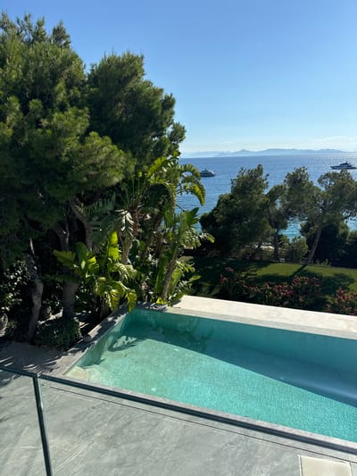 A high-angle, wide shot captures a private swimming pool overlooking the sea, likely in Vouliagmeni, Greece, given the typical architecture and landscape. The pool, rectangular and made of concrete with a pale turquoise interior, has a shallow end with steps. Its water is clear and calm, reflecting the bright blue sky. A modern glass railing with a dark metal frame runs along the foreground, bordering a tiled patio.
Beyond the pool, dense green foliage dominates the midground, featuring pine trees, large-leafed tropical plants, and bushes. The greenery creates a natural screen, offering privacy and a lush aesthetic. In the distance, the deep blue sea stretches out to the horizon, dotted with several yachts. Mountains form a hazy silhouette against the sky, suggesting a coastal mountainous region. The sky is a clear, cloudless blue, indicating a sunny day. The overall scene evokes a sense of luxury, tranquility, and a Mediterranean lifestyle.
