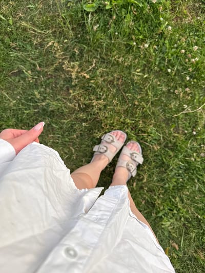This is a first-person perspective photo taken outdoors, showing a person's legs and feet from the knees down, standing on a patch of green grass. The person is wearing white two-strap sandals with gold buckles and a white shirt or dress. The grass is well-maintained and has some small white flowers scattered throughout. The lighting suggests it is daytime, likely sunny. The overall impression is casual and relaxed, possibly indicating a person enjoying a walk or relaxing in a park or garden. There is no visible text in the image. The location context provided is Ivatsevichy, Belarus.