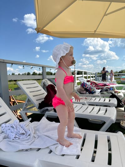 The image shows a baby lying on a white lounge chair outdoors, under a large, light-colored umbrella. The baby is wearing a white lace bonnet and a bright pink swimsuit. A pacifier is in the baby's mouth, and a beaded string with wooden beads and pacifier clips hangs around the baby's neck. The baby's legs are extended, and their feet are resting on a white towel.
In the background, several other white lounge chairs are visible, some with towels and clothing draped over them. There are also other people in the background, standing and sitting on what appears to be a deck or balcony. The scene is set against a bright blue sky with fluffy white clouds. Trees and a body of water can be seen in the distance, suggesting a waterside or park location. String lights are also visible hanging on a wooden structure.
The overall impression is a sunny, relaxed day, likely during warm weather, possibly at a resort or recreational area. The time of day appears to be daytime, with bright sunlight. No visible text is present in the image. The location context is Koloksha, Russia.