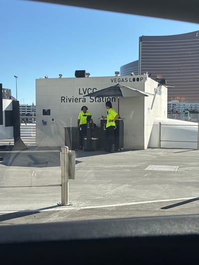 This image depicts an outdoor scene under bright daylight with a clear blue sky, located in Winchester, United States of America. The primary subject is a modern, light-colored building with a flat roof, displaying "LVCC Riviera Station" in large black lettering and "VEGAS LOOP" above it. Speakers are mounted on the roof, and security cameras are visible.

Two individuals are standing near a dark counter positioned adjacent to the building. Both are wearing bright yellow-green short-sleeved collared shirts and dark pants. The person on the left has a darker complexion and is wearing a dark headset, standing behind a dark console or stand with what appears to be a screen. They are looking towards the right. The person on the right has lighter skin, dark, medium-length hair, and is standing with one hand on their hip, looking towards the left, potentially interacting with the first individual. A large, open black market umbrella is positioned between the two individuals and the building, providing shade.

In the immediate foreground, part of a silver metal railing and a vertical post are visible, suggesting a barrier or entrance. The ground is paved with asphalt or concrete, showing some white markings. To the far left, a structure with black and white stripes and another metal barrier are visible, along with what appears to be a multi-story parking garage in the distance. In the background, behind the main building, a tall, multi-story skyscraper with a distinctive curved, reddish-brown facade and numerous horizontal lines across its surface stands prominently against the sky. Other distant buildings are also discernible.