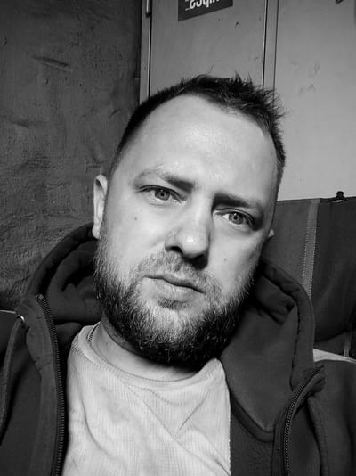 A black and white, close-up, frontal, waist-up selfie of a Caucasian male with short, dark, spiky hair and a short, full beard. He is looking directly at the camera with a neutral expression. He is wearing a light-colored, ribbed t-shirt and a dark hooded jacket that is partially zipped. The background is indistinct, with a textured, dark wall on the left and a lighter, patterned surface on the right. There is some text visible on the wall in the upper part of the frame. Based on the visible text on the wall which appears to be a Cyrillic advertisement for a tire shop, the location is likely in Salzgitter, Germany, potentially near an industrial area.