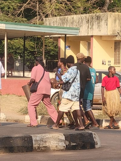 The image depicts an outdoor street scene in what is stated to be San Martín, Colombia, under bright daylight. Six individuals are walking from left to right across the foreground on an asphalt road with a weathered concrete curb painted black and white.
The leftmost walker is a man with long dark hair, dressed in a pink long-sleeved tunic and matching trousers, wearing flip-flops. He carries a black shoulder bag and a dark, cylindrical object under his right arm. Following him, partially obscured, is a person in a patterned garment. Next is a man wearing a black beanie, a light blue floral short-sleeved shirt, khaki shorts, and flip-flops, carrying a black checkered shoulder bag. Behind him, a man in a black beanie, black t-shirt, and dark shorts is visible. Another man in a teal basketball jersey (with '1' visible) and denim shorts follows. The rightmost walker is a man wearing a red short-sleeved shirt, a natural fiber skirt, and black slides.
In the background, a structure with a green sloped roof and white supports covers an area where a woman is seated. To the right, a light yellow building with brown trim, a gridded window, and posters on its wall is visible. Green trees and foliage are present throughout the background. A silver car is partially visible behind the walking figures. The environment is semi-urban with visible infrastructure and natural elements.