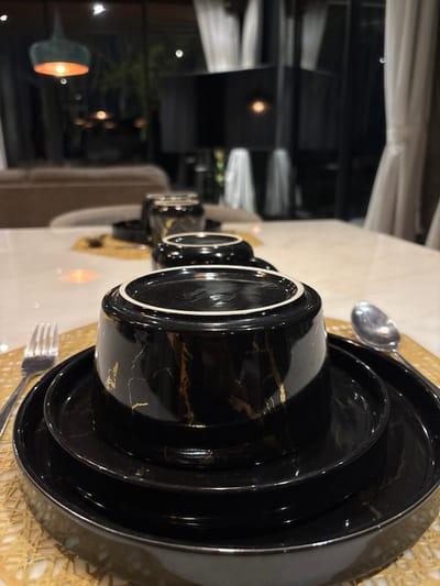 The scene depicts an interior dining setting in Batuan, Indonesia, characterized by a contemporary aesthetic. In the foreground, a stack of black ceramic dinnerware with gold marble-effect veining is centered on a light-colored, possibly golden, woven placemat. The top item is an inverted bowl or deep cup, displaying its white-rimmed base. Below it are two black plates of varying diameters, also with gold marbling. To the immediate left of the dinnerware stack, a silver fork rests on the placemat. To the immediate right, a silver spoon is positioned on the placemat.

The dinnerware is arranged on a smooth, light-colored dining table surface. Further along the table in the midground, additional out-of-focus black and gold dinnerware items, including another inverted cup and a cylindrical container, are visible. The background reveals a blurred indoor living space, featuring a light brown sofa on the left and large windows or glass doors partially covered by white curtains on the right. Multiple light sources are present, including a teal-colored pendant lamp with an orange glow on the far left, and other diffused lights throughout the dimly lit room, suggesting evening or nighttime. No individuals are present, and no actions or interactions are depicted.