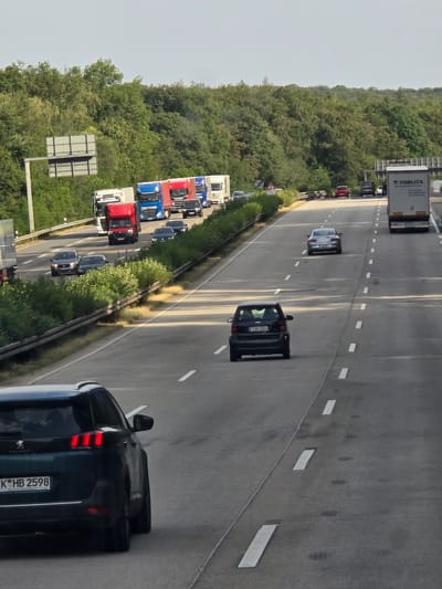The image depicts a highway scene in Bremthal, Germany, with multiple vehicles, including trucks and cars, traveling in the same direction. The highway has multiple lanes, marked by white lines. Dense green trees line the sides of the highway, creating a natural backdrop. The sky is clear and overcast.
In the foreground, a dark-colored SUV is visible, its rear facing the camera. It has a license plate that reads "K-HB 2598". Further ahead on the highway, a black Smart car is seen with its license plate reading "F-AN 2004". A silver sedan is also visible, as well as several trucks, some of which are red and blue. A white truck with the logo "TOSLITA" is in the right lane. Traffic appears to be moderate. There are no visible pedestrians.
