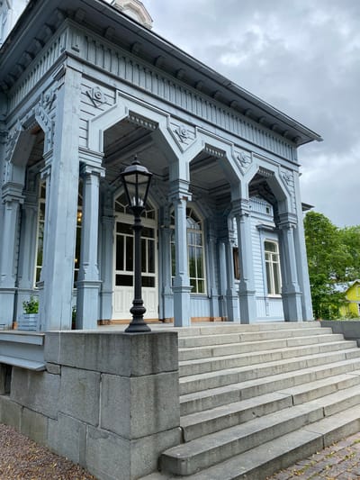 The image shows a light blue wooden building with intricate decorative elements, including carved trim and arched doorways. A wide, stone staircase leads up to the entrance, flanked by large stone blocks on the left. A black lamppost stands prominently on the staircase in front of the entrance. The building has large windows and a white double door. To the right of the building, green trees are visible, and a yellow building can be seen in the distance. The sky is overcast. The building is likely the Vyborg Architectural and Ethnographic Museum-Reserve located in Sortavala, Russia, which was once part of Finland.