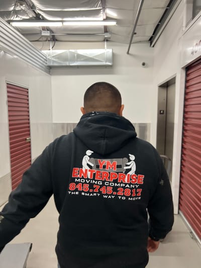 The image shows a man wearing a black hoodie with the logo of "YM ENTERPRISE MOVING COMPANY 845-745-2817 THE SMART WAY TO MOVE" on the back. He is standing in what appears to be a storage unit facility. The facility has red storage unit doors and white walls. The location is likely in Town of New Windsor, United States of America, based on the area code in the phone number on the hoodie.