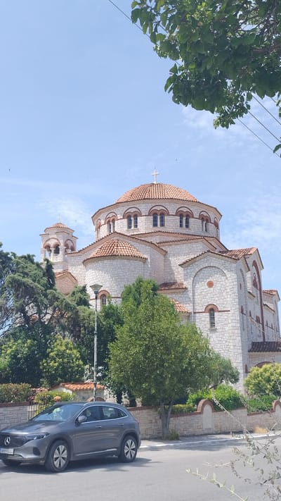 In Νέα Μηχανιώνα, Greece, a grey electric car is parked on the side of a street in front of a large, stone-built church with red tile roofs. The church has multiple domes and bell towers, and is adorned with arched windows. Lush green trees and a stone wall with decorative elements line the street between the car and the church. The sky above is a clear blue with a few wispy clouds. Overhead, tree branches with green leaves are visible, along with power lines. A street lamp is positioned near the church. The car's license plate reads "XHY-1464". No people are prominently visible in the scene.