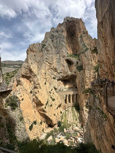 The image depicts a view of the Caminito del Rey, a walkway pinned along the steep walls of a narrow gorge in El Chorro, near Ardales in the province of Málaga, Spain. The walkway appears to be built into the sheer rock face, with a group of people visible walking on the platform above and below. The walkway follows the contours of the canyon with vegetation growing on the rock face. At the bottom of the gorge a river flows over rocks. The sky is visible above the canyon and clouds can be seen. A building with 3 windows is also seen in the image.